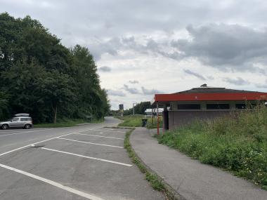 Leerer Parkstreifen mit 6 Markierungen für die Schrägaufstellung von PKW. Daneben ein Gehweg und ein gemauertes WC-Gebäude mit rotem Dach.