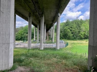 Das Bild zeigt den Blick aus Richtung Widerlager unter die Talbrücke auf die Brückenpfeiler und die Regenwasserbehandlungsanlage. Umliegend ist Grünland zu erkennen sowie Wald im Hintergrund des Bildes.