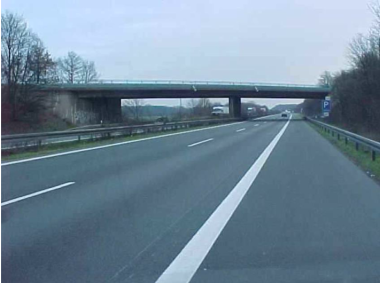 Sicht vom Standstreifen einer Fahrtrichtung der Bundesautobahn 1 auf die Queransicht des Brückenbauwerks Schmedehausener Straße mit beidseitig flankierender Gehölzstruktur entlang der Bundesautobahn.