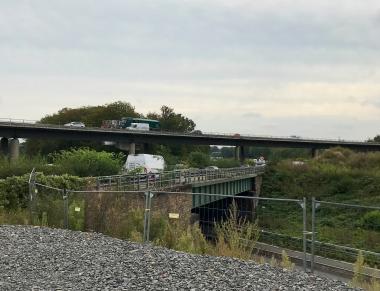 Das Bild zeigt zwei Autobahnbrücken auf denen viele Fahrzeuge fahren. Eine Brücke hat mehrere Betonpfeiler. Im Vordergrund des Bildes ist ein Bauzaun zu sehen. 