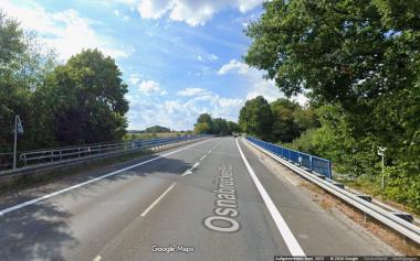 Auf dem Bild ist die zweisprurige Osnabrücker Straße, die die Bundesautobahn 1 kreuzt, zu sehen. Die Blickrichtung ist gen Westen. Der Straßenrand weist Gehözstrukturen auf. Die eigentliche Überführung über die Bundesautobahn ist mit einem Geländer in blauer Farbe begrenzt.